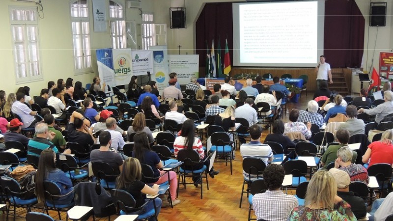 Público presente no evento, de frente para o palco. À esquerda, ao lado do palco, banners das instituições promotoras do Seminário seguidos de quatro bandeiras. Ao centro, um telão com o conteúdo de uma das palestras e, à direita, um palestrante em pé.