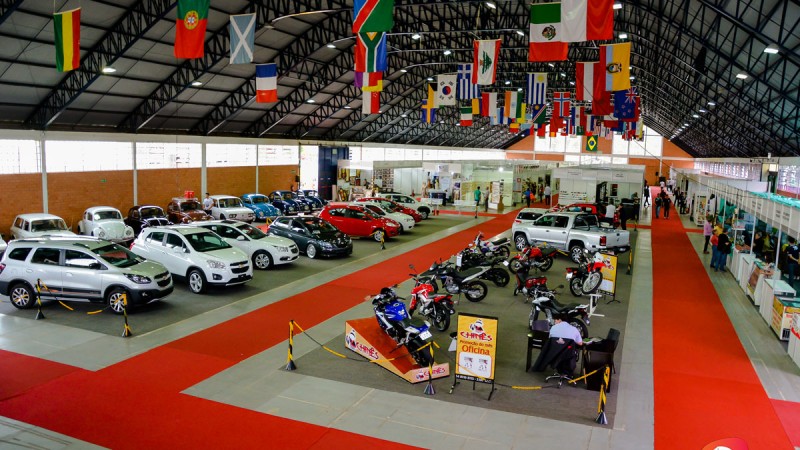 Pavilhão da Feira com expositores. Corredor sinalizado por tapetes laranja. À esquerda, exposição de carros, ao centro motos e, à direita, estandes dos demais expositores. No teto, bandeiras de diversos países penduradas.