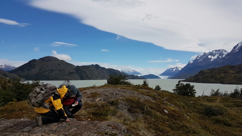 Duas pessoas estão agachadas sobre uma rocha coletando musgos. Ao fundo, montanhas na Patagônia e oceano Atlântico.