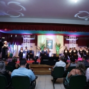 Em um palco decorado, uma mulher de toga branca fala ao microfone em um púlpito e outras dezoito assistem sentadas, vestindo togas pretas e azuis.