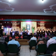 Em um palco decorado, uma mulher de toga branca fala ao microfone em um púlpito e outras dezoito assistem sentadas, vestindo togas pretas e azuis.