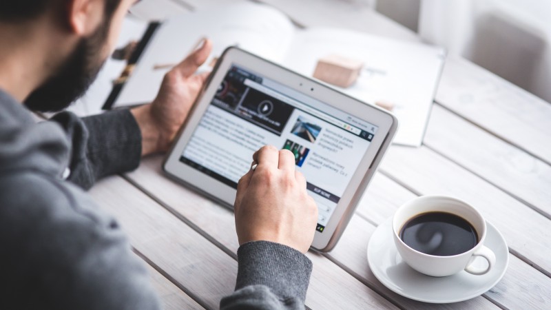 Homem segura um tablet e seleciona notícias em um site enquanto apoia o equipamento sobre uma mesa, assim como uma xícara de café e uma revista aberta.