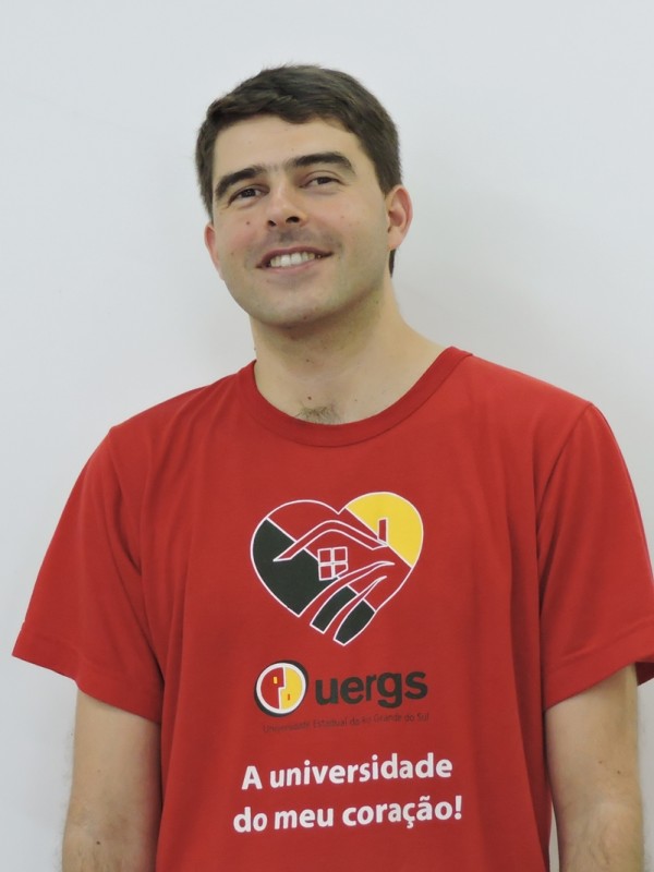 Homem de sorridente de pele clara e cabelos castanhos e curtos. Veste camiseta vermelha com o logotipo da Uergs.