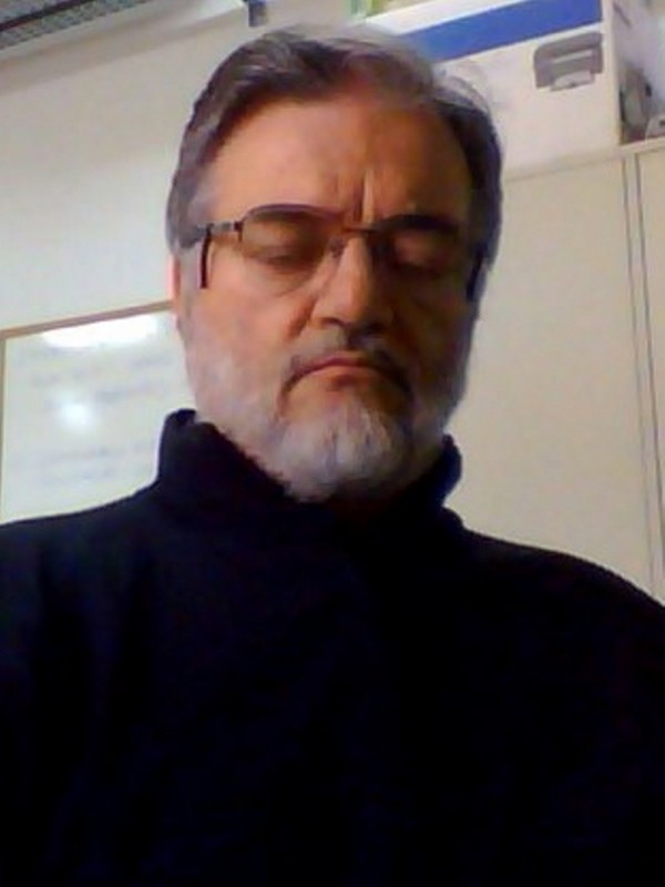 Homem de pele clara, cabelo, barba e bigode grisalhos. Usa óculos de grau, veste blusa de gola alta preta e está com semblante sério e de olhos fechados.