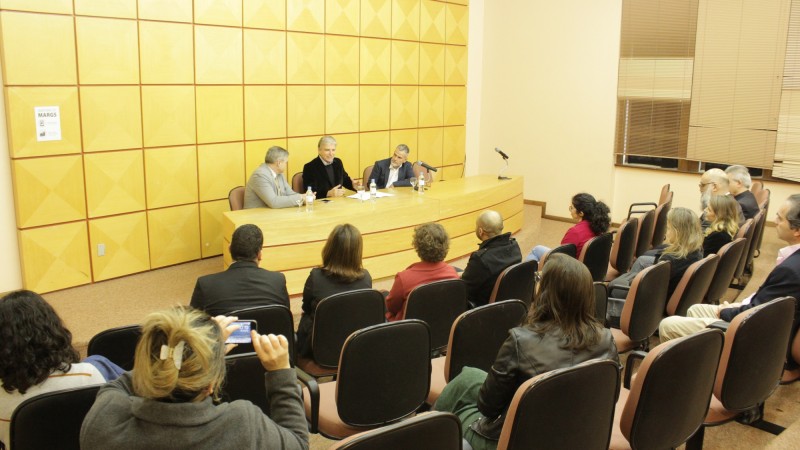 Sentados à mesa em um auditório, estão o secretário Luís Lamb, o presidente da Fundação Bienal do Mercosul, Gilberto Schwartsmann, e o reitor da Uergs, Leonardo Beroldt. Em frente a eles, a plateia que assiste o ato.