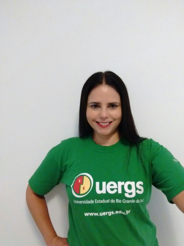 Mulher sorridente de pele morena, cabelos pretos, lisos e compridos. Usa camiseta verde com o logotipo da Uergs.