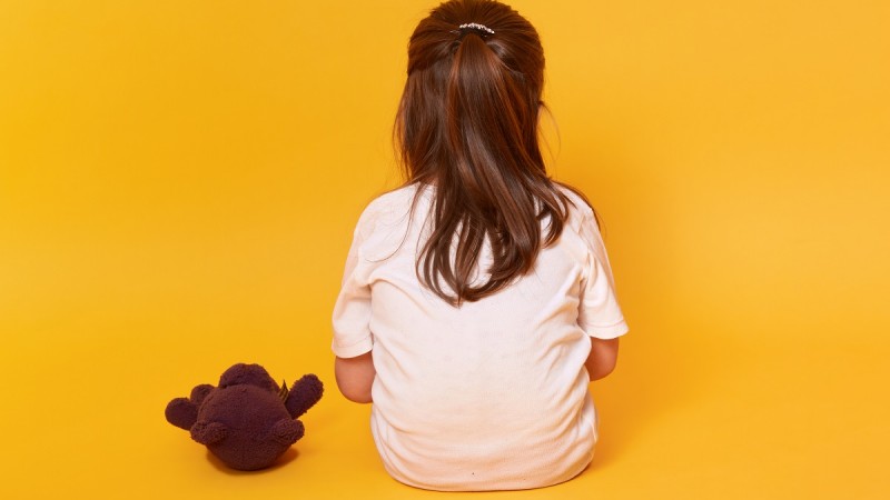 Foto colorida. Fundo amarelo. Ao centro, menina sentada de costas. Ao lado esquerdo, pequeno urso de pelúcia marrom.