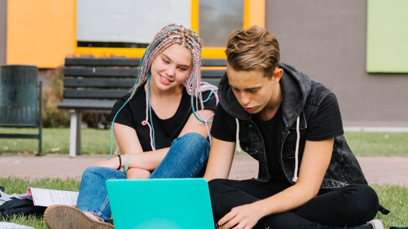 Um casal de adolescentes sentados em um gramado utilizam um notebook.