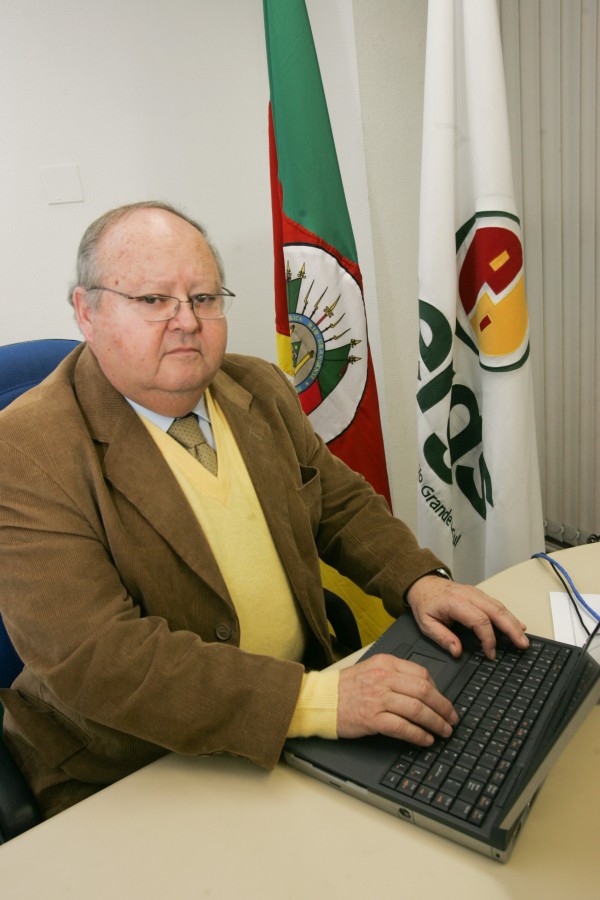 Foto colorida. Homem branco, de meia-idade, usa terno marrom, colete amarelo, gravata e camisa. Ele está sentado e digita em um notebook. ao fundo, bandeiras da Uergs e Rio Grande do Sul.