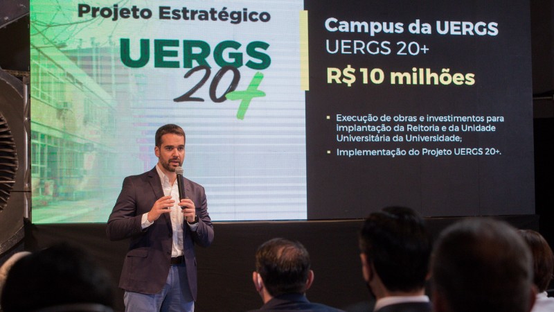 Foto colorida do governador gaúcho Eduardo Leite de pé em frente a uma tela em que se lê Projeto Estratégico UERGS 20+; Campus da UERGS R$ 10 milhões.