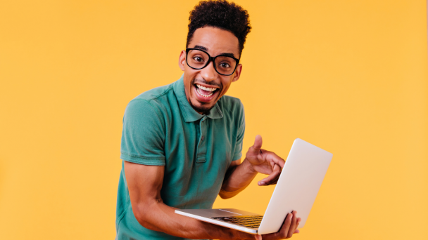 Foto colorida. Sobre fundo amarelo, jovem negro, de óculos de grau, sorridente, está em pé, apoiando um notebook com o braço direito.