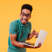 Foto colorida. Sobre fundo amarelo, jovem negro, de óculos de grau, sorridente, está em pé, apoiando um notebook com o braço direito.