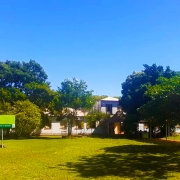 Amplo gramado à frente do prédio da Unidade, que está atrás de várias árvores. à esquerda, placa de identificação.