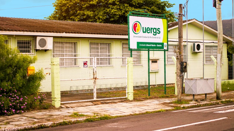 Foto do prédio da Unidade, de apenas 1 andar e de paredes em tom de verde claro. À frente, junto à cerca, a placa de identificação da Uergs.