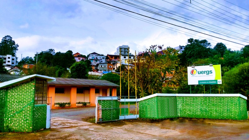 Em primeiro plano, um muro de tijolos à vista pintados de verde, com uma placa de identificação da Uergs, à direita. Ao fundo, o prédio da Unidade, pintado de laranja.