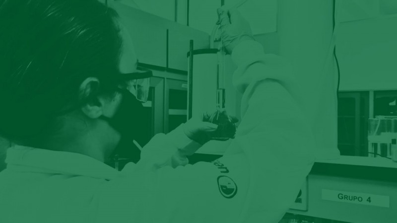 Foto com filtro verde de uma mulher operando um equipamento de laboratório. Ela usa luvas e um jaleco de mangas longas com o logo da Uergs bordado na manga direita.