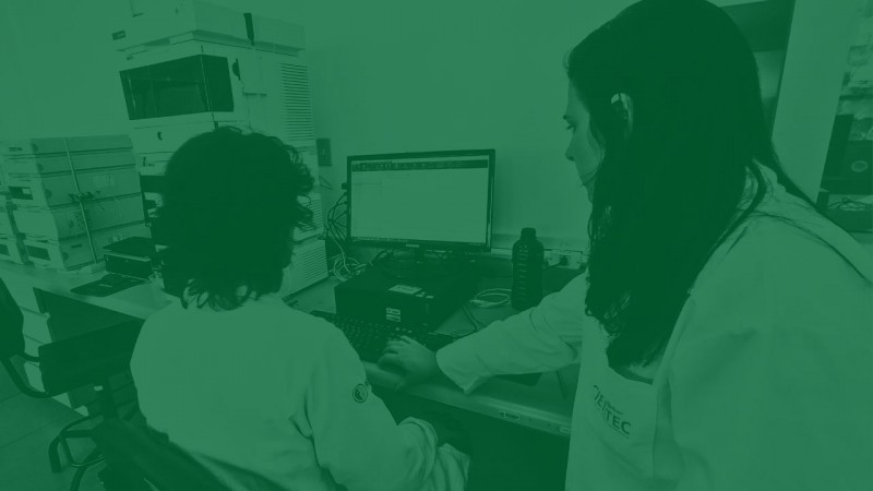 Foto com filtro verde de um homem de cabelos volumosos visto de costas, sentado em frente a um computador e a um cromatógrafo. Ao seu lado, uma mulher de cabelos longos lhe explica algo.
