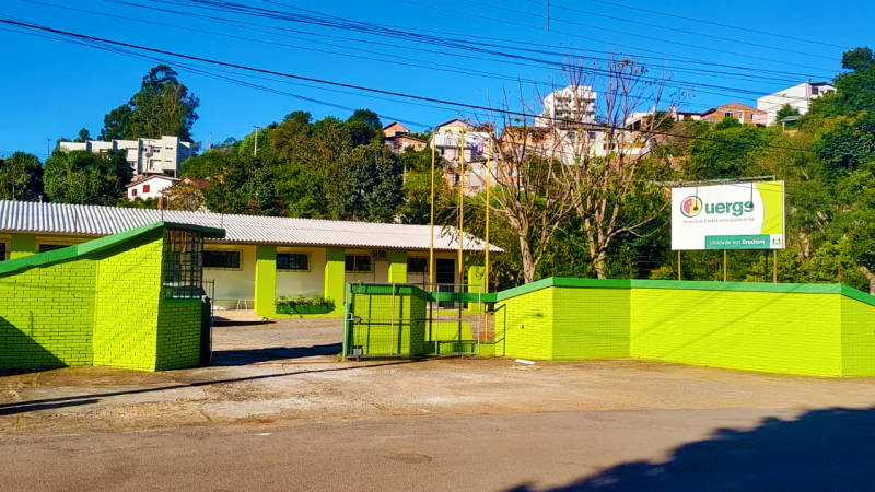 Em primeiro plano, um muro de tijolos à vista pintados de verde-limão, com uma placa de identificação da Uergs, à direita. Ao fundo, o prédio da Unidade, pintado de tom claro com vigas em verde-limão.