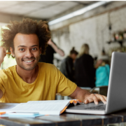 Um jovem negro de pele clara, com cabelo crespo volumoso, sorridente, sentado com livros e um notebook à sua frente.