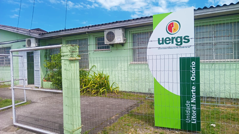 Foto colorida de uma construção de apenas um andar, com as paredes pintadas de verde claro, grades na janela e uma cerca de aço. Dentro do pátio, há um totem em tons de verde com o logo da Uergs, escrito Unidade Litoral Norte - Osório.