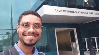 Foto de Leonardo Pinheiro, um homem negro, sorrindo e usando óculos com armação fina e arredondada. Ele está em frente ao Ministério da Pesca e Aquicultura.