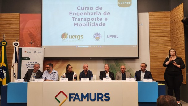 Foto colorida de pessoas sentadas atrás de uma mesa. Na mesa, lê-se Famurs. São sete pessoas sentadas. Elas são as autoridades do evento. Atrás e acima delas, há um telão com a identidade visual do evento.