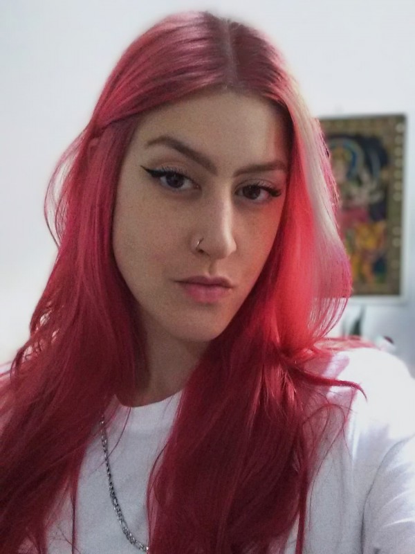 Foto colorida. Uma jovem de pele clara e olhos escuros, com cabelos longos vermelhos, vestindo camiseta branca.