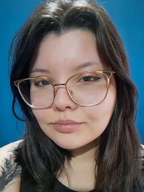 Foto colorida. Uma jovem de pele clara, olhos escuros e cabelo médio na cor castanho escuro, usa óculos de grau com aros dourados.