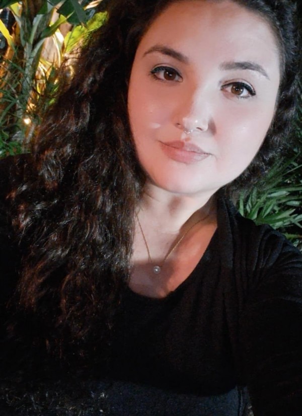 Foto de uma jovem de pele clara, cabelos longos cacheados escuros, olhos escuros, veste blusa preta.