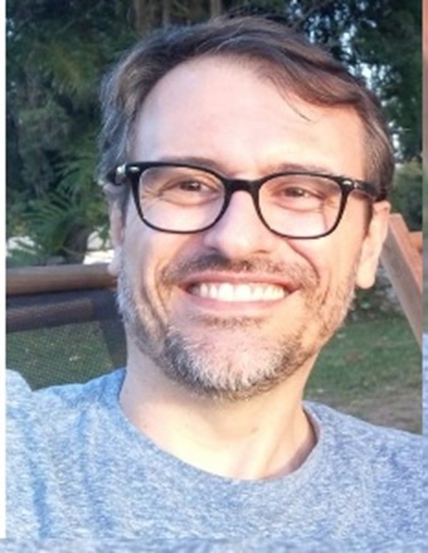 Homem de pele clara, com cabelos e barbas claros e grisalhos, de olhos escuros; usa óculos de grau com aros pretos e está sorridente.
