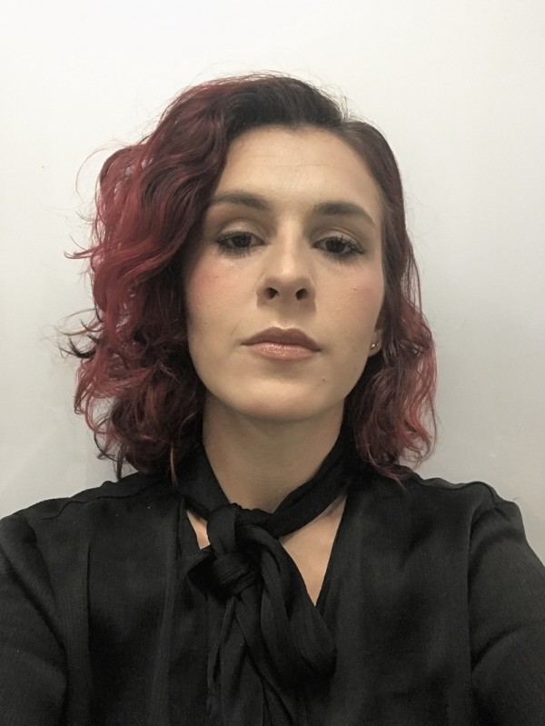 Jovem de pele clara, olhos escuros, cabelos curtos castanhos com mechas vermelhas nas pontas; veste camisa preta com laço no pescoço.
