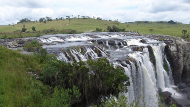 A imagem mostra uma grande cachoeira de queda larga e pouco inclinada, localizada em uma região de campo aberto. A água escorre em diversos filetes sobre uma formação rochosa escura, formando várias pequenas quedas que se encontram em uma queda principal mais alta, à direita da imagem. A vegetação ao redor é predominante de gramíneas e arbustos, típica dos campos sulinos. Algumas árvores isoladas, como araucárias, aparecem ao fundo, em um terreno levemente ondulado. O céu está parcialmente nublado, com nuvens brancas e cinzentas cobrindo grande parte do azul.