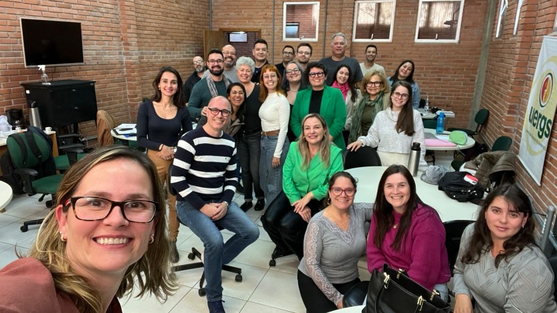 Foto colorida tirada em ambiente interno, numa sala com paredes de tijolos à vista e três janelas pequenas na parte superior. O espaço tem piso branco e é mobiliado com cadeiras giratórias e mesas redondas brancas.
No centro da imagem, há um grupo de aproximadamente 25 pessoas, adultas, a maioria aparentando estar entre os 30 e 60 anos, posando sorridentes para a foto. Estão vestidas de forma casual, com roupas em tons variados como verde, cinza, rosa, preto, branco e azul.
No canto esquerdo inferior da imagem, uma mulher de óculos e cabelos loiros escuros na altura dos ombros tira a selfie. Ela está sorrindo e veste uma blusa marrom.
Ao fundo, à direita, há uma faixa com o logotipo da Uergs.