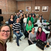 Foto colorida tirada em ambiente interno, numa sala com paredes de tijolos à vista e três janelas pequenas na parte superior. O espaço tem piso branco e é mobiliado com cadeiras giratórias e mesas redondas brancas.
No centro da imagem, há um grupo de aproximadamente 25 pessoas, adultas, a maioria aparentando estar entre os 30 e 60 anos, posando sorridentes para a foto. Estão vestidas de forma casual, com roupas em tons variados como verde, cinza, rosa, preto, branco e azul. 
No canto esquerdo inferior da imagem, uma mulher de óculos e cabelos loiros escuros na altura dos ombros tira a selfie. Ela está sorrindo e veste uma blusa marrom.
Ao fundo, à direita, há uma faixa com o logotipo da Uergs.
