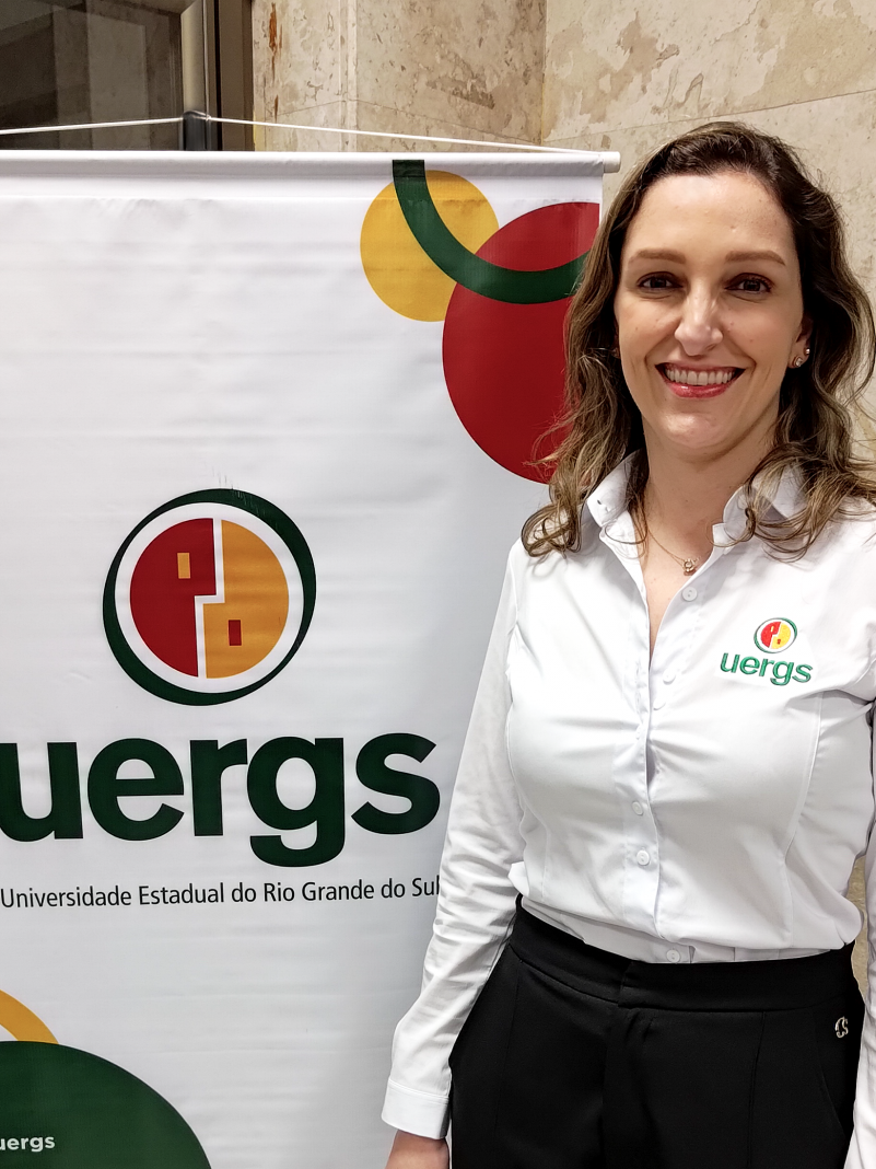 Mulher sorridente de pele cara, olhos castanhos e cabelo louro, de tamanho médio e levemente ondulado. Veste camisa branca, com logotipo da Uergs bordado, localizado no lado esquerdo do peito, e calça preta. Atrás dela, um banner com o logotipo da Uergs, com fundo branco.