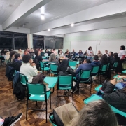 A foto mostra uma sala de aula ampla e bem iluminada, com piso de madeira e paredes de tijolos pintados de verde claro. Cerca de 60 pessoas participam de uma roda de conversa, dispostas em cadeiras escolares com mesas acopladas, organizadas em formato circular e semicircular. A maioria das cadeiras é verde água.
No centro da sala, duas mulheres de camiseta branca falam para o grupo. Uma está em pé, próximo ao quadro branco, e outra ao centro da roda, segurando um leque com a mesma identidade visual da camiseta.
O público é diverso, com pessoas de diferentes faixas etárias, tons de pele, estilos e gêneros. Algumas estão atentas às falas, outras leem folhetos ou observam ao redor. Quase todas carregam o leque colorido da campanha sobre igualdade.
Ao fundo, grandes janelas com vidros escuros ocupam a maior parte da parede lateral. 
