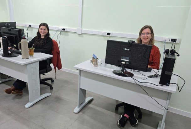 Duas mulheres estão sentadas em frente a mesas de escritório, posicionadas lado a lado em um ambiente interno com paredes de cor clara. Ambas sorriem para a câmera. À esquerda da imagem, a mulher tem cabelos longos e escuros, veste um suéter preto e calça escura, e usa botas marrons. Sobre a mesa dela há um computador, uma garrafa térmica e uma cuia com chimarrão. Um casaco vermelho está pendurado na parte de trás da cadeira.
À direita da imagem, a outra mulher tem cabelos loiros na altura dos ombros e usa óculos. Ela veste uma blusa vermelha escura e calça preta, com tênis pretos. Sobre a mesa dela há um computador, uma caneca branca e um calendário. O chão é de cor cinza e o ambiente parece iluminado por luz artificial.
