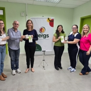 Seis pessoas estão em pé, lado a lado, sorrindo para a câmera em um ambiente interno com paredes verde-claro e portas verdes. No centro do grupo, ao fundo, há um banner com o logotipo da Uergs (Universidade Estadual do Rio Grande do Sul).
Da esquerda para a direita:
A primeira pessoa é uma mulher de cabelos castanhos, soltos e ondulados, usando óculos, blusa branca de mangas bufantes e jeans. Ela está com uma das mãos no ombro do homem ao lado.


O segundo é um homem de cabelo grisalho curto, usando óculos, camisa azul-clara listrada e calça jeans. Ele segura uma caneca branca junto com a terceira pessoa.


A terceira é uma mulher loira, de óculos, usando blusa azul-escura e calça preta. Ela segura a caneca e um panfleto.


A quarta é uma mulher de cabelos castanhos escuros e longos, usando uma blusa verde e calça preta. Ela segura o mesmo tipo de panfleto.


A quinta é uma mulher de óculos com armação azul, cabelos presos, vestindo uma camiseta azul com branco e calça esportiva. Ela segura um copo transparente.


A sexta é uma mulher de cabelos loiros, soltos, vestindo uma blusa rosa-choque e calça jeans escura. Ela está com a mão nas costas da colega ao lado.


O ambiente é iluminado por luz artificial, com piso cinza claro e teto de placas modulares. Todos parecem felizes e participando de um momento comemorativo ou de entrega simbólica de brindes.
