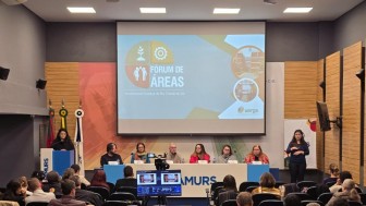Fotografia colorida tirada em um auditório durante a abertura do Fórum de Áreas. No centro da imagem, há um painel com sete pessoas sentadas atrás de uma longa mesa branca, sobre um tablado. Eles estão participando da mesa de abertura. Na frente da mesa está o logotipo da Famurs (Federação das Associações de Municípios do Rio Grande do Sul), local onde o evento está sendo realizado.
Acima dos participantes, há uma grande tela de projeção com a identidade visual do evento: o título “Fórum de Áreas”, seguido da inscrição “Universidade Estadual do Rio Grande do Sul”. O fundo do slide tem formas geométricas coloridas e ícones que representam pessoas, uma planta e uma engrenagem.
À esquerda da imagem, uma mulher veste preto e fala ao microfone em um púlpito azul, também com a marca da Famurs. Atrás dela, três bandeiras estão posicionadas, entre elas a do Brasil, do Rio Grande do Sul e da Famurs.
À direita, em frente à parede lateral do auditório, uma mulher de cabelo castanho escuro e longo faz interpretação em Língua Brasileira de Sinais (Libras). O público aparece de costas, sentado em cadeiras organizadas em fileiras. Em primeiro plano, vê-se a equipe de filmagem, com dois monitores exibindo a transmissão ao vivo do evento.
O ambiente é bem iluminado, com paredes em tons neutros e painéis de madeira nas laterais. 
