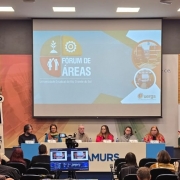 Fotografia colorida tirada em um auditório durante a abertura do Fórum de Áreas. No centro da imagem, há um painel com sete pessoas sentadas atrás de uma longa mesa branca, sobre um tablado. Eles estão participando da mesa de abertura. Na frente da mesa está o logotipo da Famurs (Federação das Associações de Municípios do Rio Grande do Sul), local onde o evento está sendo realizado.
Acima dos participantes, há uma grande tela de projeção com a identidade visual do evento: o título “Fórum de Áreas”, seguido da inscrição “Universidade Estadual do Rio Grande do Sul”. O fundo do slide tem formas geométricas coloridas e ícones que representam pessoas, uma planta e uma engrenagem.
À esquerda da imagem, uma mulher veste preto e fala ao microfone em um púlpito azul, também com a marca da Famurs. Atrás dela, três bandeiras estão posicionadas, entre elas a do Brasil, do Rio Grande do Sul e da Famurs.
À direita, em frente à parede lateral do auditório, uma mulher de cabelo castanho escuro e longo faz interpretação em Língua Brasileira de Sinais (Libras). O público aparece de costas, sentado em cadeiras organizadas em fileiras. Em primeiro plano, vê-se a equipe de filmagem, com dois monitores exibindo a transmissão ao vivo do evento.
O ambiente é bem iluminado, com paredes em tons neutros e painéis de madeira nas laterais. 
