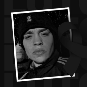Foto em preto e branco de um jovem sério, moreno claro, de sobrancelhas e lábios espessos, vestindo jaqueta e uma touca de lã pretas. Sobre a foto, há a imagem de um laço preto, simbolizando luto.