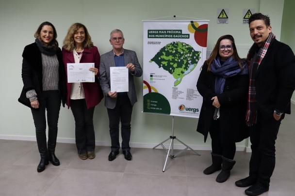 Cinco pessoas estão em pé lado a lado, posando para a foto em ambiente interno, sobre piso claro e parede verde-clara lisa. Ao centro da imagem, uma mulher de cabelos loiros ondulados e um homem de cabelos curtos grisalhos seguram documentos à frente do corpo, sorrindo para a câmera. À esquerda, outra mulher de cabelos castanhos ondulados, trajando sobretudo preto e cachecol escuro, também sorri. À direita da dupla central, uma mulher de cabelos longos castanhos escuros, com óculos de armação azul, usa casaco preto e cachecol azul marinho. Ao lado dela, um homem de bigode e cavanhaque veste sobretudo escuro e cachecol xadrez. À direita do grupo, há um banner com o título “UERGS MAIS PRÓXIMA DOS MUNICÍPIOS GAÚCHOS” e um mapa do Rio Grande do Sul com a localização das unidades da Universidade. Ao fundo, placas de aviso de risco elétrico estão fixadas na parede. 