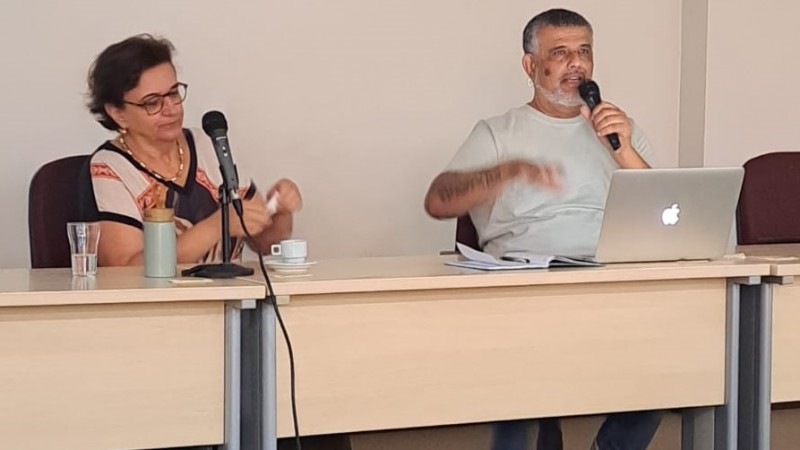 Fotografia de duas pessoas sentadas lado a lado atr&aacute;s de uma mesa, participando de uma atividade em ambiente fechado. &Agrave; esquerda, uma mulher de pele clara, cabelos castanhos curtos e ondulados, usa &oacute;culos e est&aacute; sorrindo levemente. Ela veste uma blusa com estampa geom&eacute;trica em tons de preto, bege e laranja. &Agrave; sua frente, sobre a mesa, h&aacute; dois copos de vidro com &aacute;gua, uma garrafa t&eacute;rmica verde com tampa de madeira, uma x&iacute;cara branca com pires e um microfone em suporte.

&Agrave; direita, um homem de pele escura, com cabelo curto grisalho e barba, segura um microfone com a m&atilde;o direita e parece estar falando. Ele veste camiseta cinza clara e jeans azul. Sobre a mesa &agrave; sua frente, h&aacute; um notebook aberto (marca Apple), algumas folhas de papel e outro microfone em suporte. Ambos est&atilde;o sentados em cadeiras de escrit&oacute;rio, atr&aacute;s de uma mesa de tampo bege claro com estrutura met&aacute;lica cinza. Ao fundo, uma parede bege clara. A imagem sugere um momento de fala ou apresenta&ccedil;&atilde;o em uma mesa de debate ou reuni&atilde;o.
