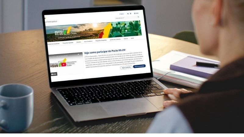 Pessoa sentada à mesa utiliza um notebook, que exibe na tela o site do “Pacto RS 25”. A página mostra o logotipo do programa, com as cores verde, amarela e vermelha, e o título “Veja como participar do Pacto RS 25!”. Ao lado do computador há uma caneca azul-clara, um caderno roxo e uma caneta.