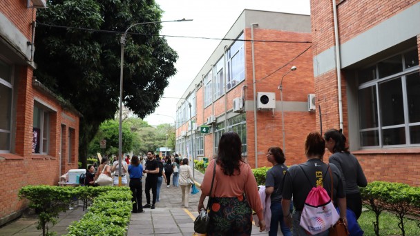 Foto horizontal. Em primeiro plano, v&ecirc;-se um corredor externo de uma universidade, com caminho pavimentado e canteiros verdes nas laterais. &Agrave; direita e &agrave; esquerda, h&aacute; pr&eacute;dios de dois andares com paredes de tijolos aparentes, janelas grandes e aparelhos de ar-condicionado fixados nas fachadas. No corredor, v&aacute;rias pessoas caminham, vistas principalmente de costas e de perfil. S&atilde;o adultos de diferentes apar&ecirc;ncias f&iacute;sicas, usando roupas casuais, mochilas e bolsas, sugerindo um ambiente acad&ecirc;mico em funcionamento. Ao fundo, o corredor segue em perspectiva, com mais pessoas e vegeta&ccedil;&atilde;o.
