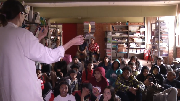 Mulher em p&eacute; &agrave; esquerda, vestindo jaleco e virada de costas, segurando uma pin&ccedil;a com um algod&atilde;o em chamas. A sua frente, est&atilde;o diversas crian&ccedil;as sentadas ao ch&atilde;o que olham a demonstra&ccedil;&atilde;o. Elas t&ecirc;m entre 9 e 11 anos e est&atilde;o em uma biblioteca.