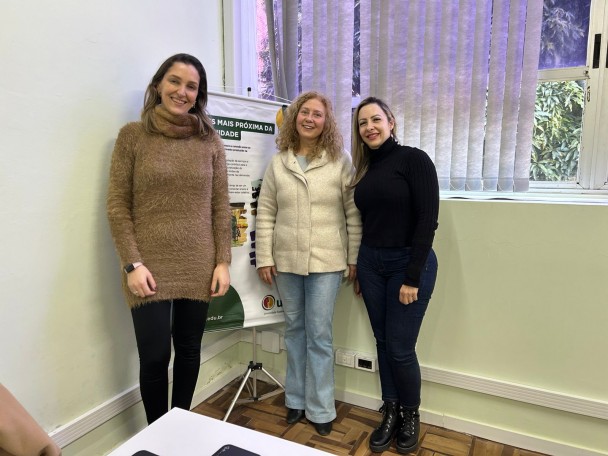 Fotografia em ambiente interno, com três mulheres em pé, lado a lado, voltadas para a câmera e sorrindo. À esquerda, uma mulher de estatura média, pele clara, cabelos castanhos longos e soltos. Ela veste um vestido de manga longa, de tecido felpudo, em tom marrom claro, calça preta e usa um relógio no pulso esquerdo. Ao centro, uma mulher de pele clara, cabelos claros e cacheados, na altura dos ombros. Ela veste um casaco claro, fechado, calça jeans azul clara e sapatos escuros. À direita, uma mulher de pele clara, cabelos castanhos longos e soltos. Ela usa blusa preta de manga longa, calça jeans escura e botas pretas.

Atrás das três, há um banner institucional apoiado em um tripé, com predominância das cores branco e verde e logotipos na parte inferior. O ambiente tem paredes claras, piso de madeira e uma janela com persianas verticais parcialmente abertas, permitindo a entrada de luz natural. Em primeiro plano, aparece parcialmente uma mesa branca.