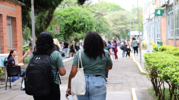 Fotografia em ambiente externo de um campus universit&aacute;rio. Em primeiro plano, duas mulheres caminham de costas por uma cal&ccedil;ada larga pavimentada com blocos de concreto. Ambas usam camisetas verdes. A mulher &agrave; esquerda tem cabelos escuros e ondulados na altura dos ombros e carrega uma mochila preta nas costas. A mulher &agrave; direita tem cabelos longos e cacheados e leva uma bolsa de tecido clara pendurada no ombro; ela tamb&eacute;m segura uma garrafa t&eacute;rmica prateada na m&atilde;o direita e veste uma saia ou cal&ccedil;a jeans clara.

Ao longo da cal&ccedil;ada, h&aacute; canteiros com arbustos baixos e bem cuidados. &Agrave; direita, aparece a fachada de um pr&eacute;dio de tijolos com janelas e uma placa verde com o n&uacute;mero &ldquo;4&rdquo;. &Agrave; esquerda, outro pr&eacute;dio de tijolos e algumas pessoas sentadas em cadeiras pr&oacute;ximas a uma mesa.

No fundo da imagem, v&aacute;rios estudantes caminham ou conversam em pequenos grupos sob &aacute;rvores grandes e frondosas, que formam uma &aacute;rea verde e sombreada t&iacute;pica de um campus universit&aacute;rio.