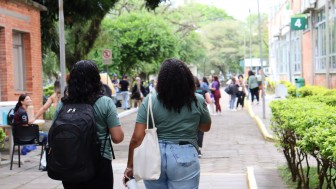 Fotografia em ambiente externo de um campus universit&aacute;rio. Em primeiro plano, duas mulheres caminham de costas por uma cal&ccedil;ada larga pavimentada com blocos de concreto. Ambas usam camisetas verdes. A mulher &agrave; esquerda tem cabelos escuros e ondulados na altura dos ombros e carrega uma mochila preta nas costas. A mulher &agrave; direita tem cabelos longos e cacheados e leva uma bolsa de tecido clara pendurada no ombro; ela tamb&eacute;m segura uma garrafa t&eacute;rmica prateada na m&atilde;o direita e veste uma saia ou cal&ccedil;a jeans clara.

Ao longo da cal&ccedil;ada, h&aacute; canteiros com arbustos baixos e bem cuidados. &Agrave; direita, aparece a fachada de um pr&eacute;dio de tijolos com janelas e uma placa verde com o n&uacute;mero &ldquo;4&rdquo;. &Agrave; esquerda, outro pr&eacute;dio de tijolos e algumas pessoas sentadas em cadeiras pr&oacute;ximas a uma mesa.

No fundo da imagem, v&aacute;rios estudantes caminham ou conversam em pequenos grupos sob &aacute;rvores grandes e frondosas, que formam uma &aacute;rea verde e sombreada t&iacute;pica de um campus universit&aacute;rio.