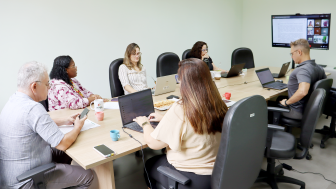 Foto de uma mesa de reuni&otilde;es com 6 pessoas sentadas, sendo 2 homens e 4 mulheres. Eles olham para dispositivos eletr&ocirc;nicos como celulares e laptops. Ao fundo, uma TV projeta a transmiss&atilde;o da sess&atilde;o, com pessoas participando virtualmente.